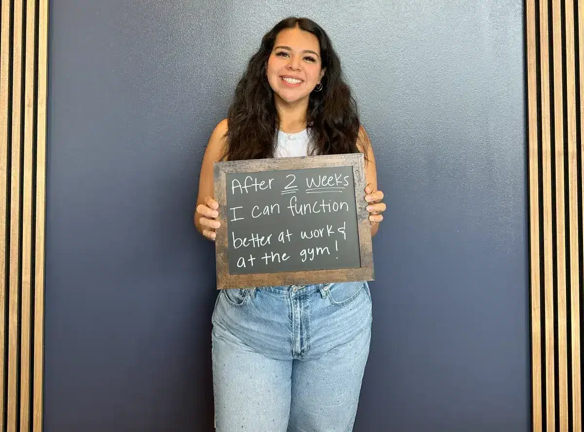 Limitless Chiropractic female patient smiling while holding a sign that says “After 2 weeks I can function better at work & at the gym!” — showing improved mobility and performance through chiropractic care in Alliance, Fort Worth.